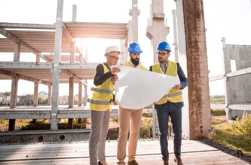 Three people wearing yellow vests and helmets are on the construction site. They look at the mockup together.