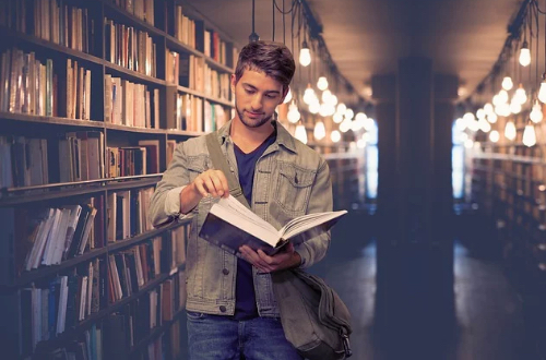Mężczyzna czyta książkę pomiędzy regałami w bibliotece.